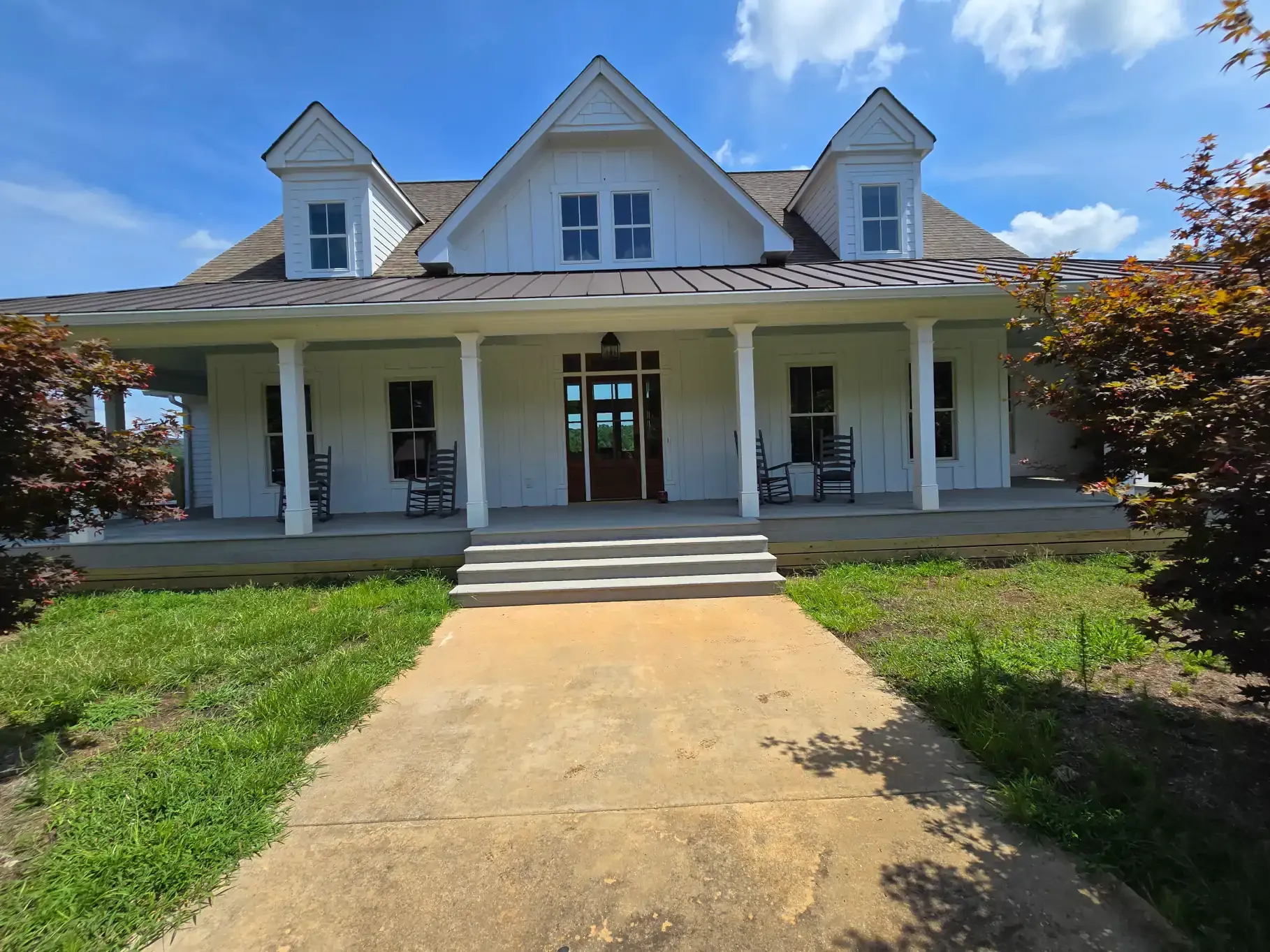A two-story white farmhouse with a large front porch and three dormers under a blue sky, approached by a concrete walkway.