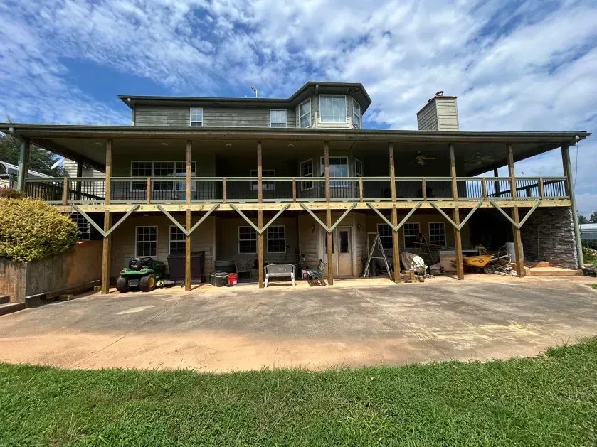 Large multi-story house with a wide wooden deck, support pillars, and a concrete patio on a grassy lawn under a blue sky.