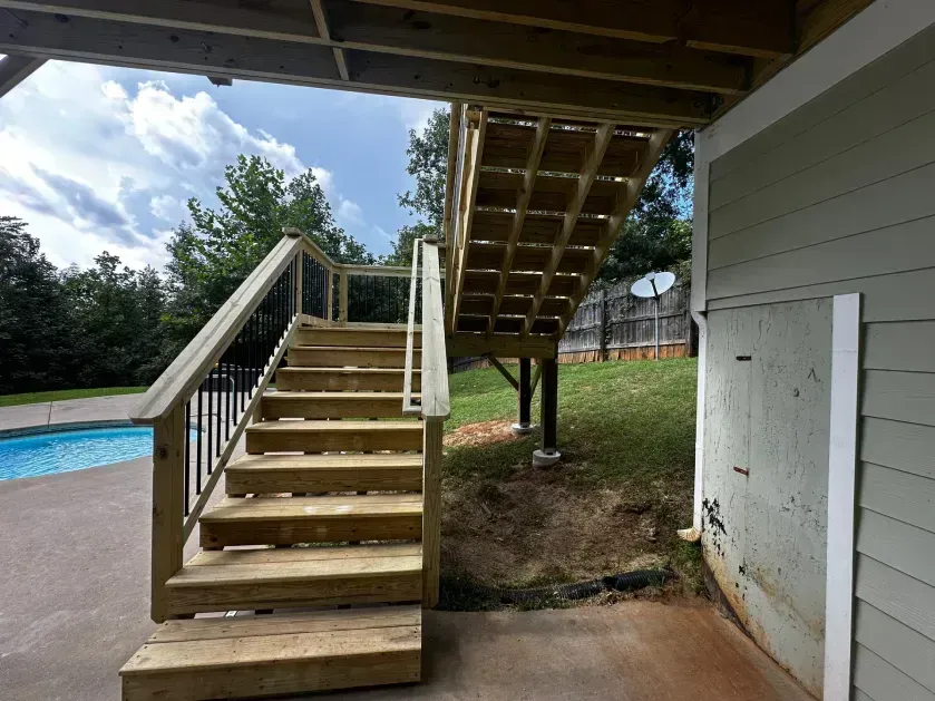 Wooden outdoor staircase leading from a concrete patio up to a deck, with a swimming pool visible in the background.