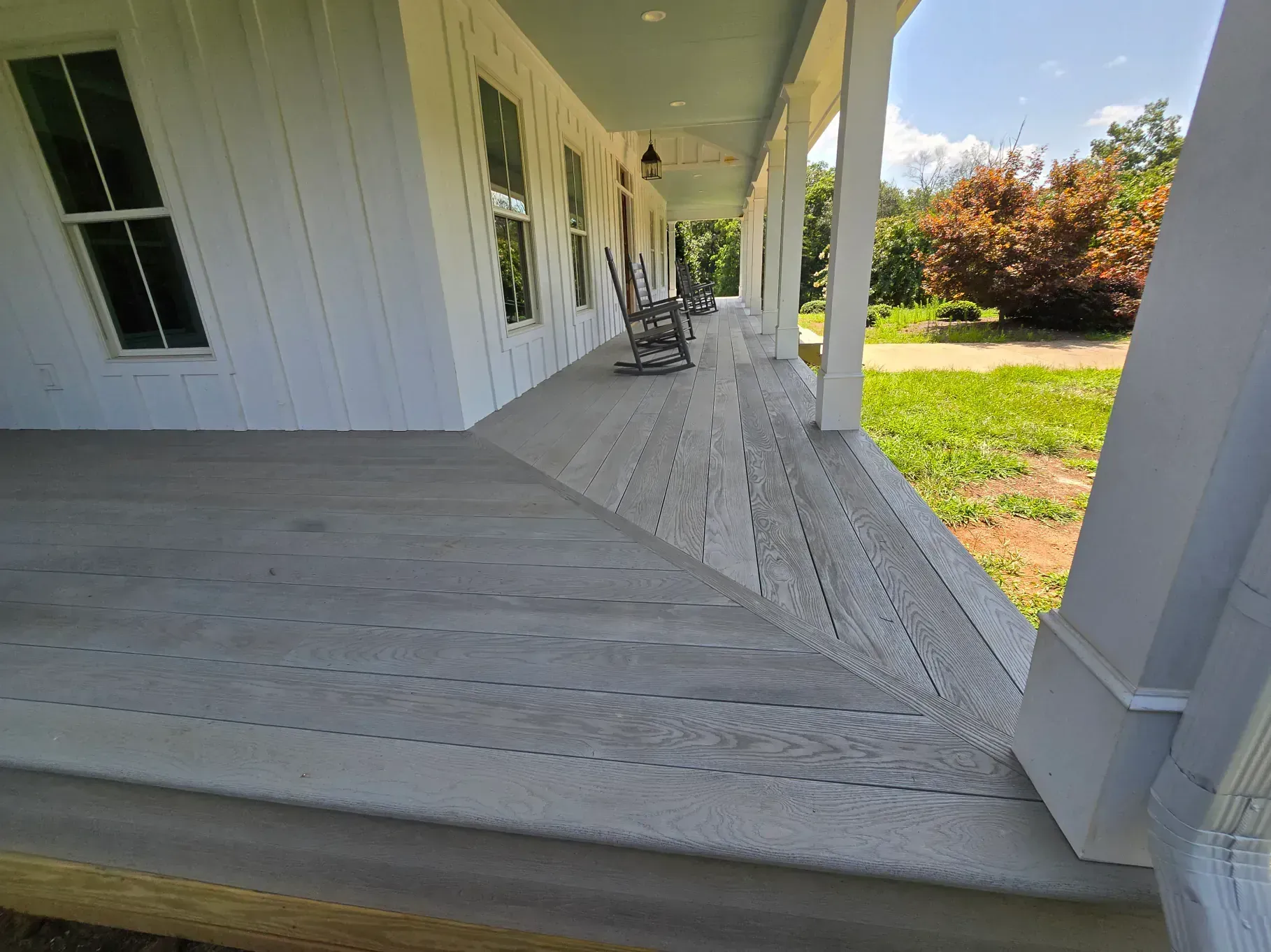 A wide, light-gray wooden porch deck with a mitered corner, overlooking a lawn with green foliage and a tree.
