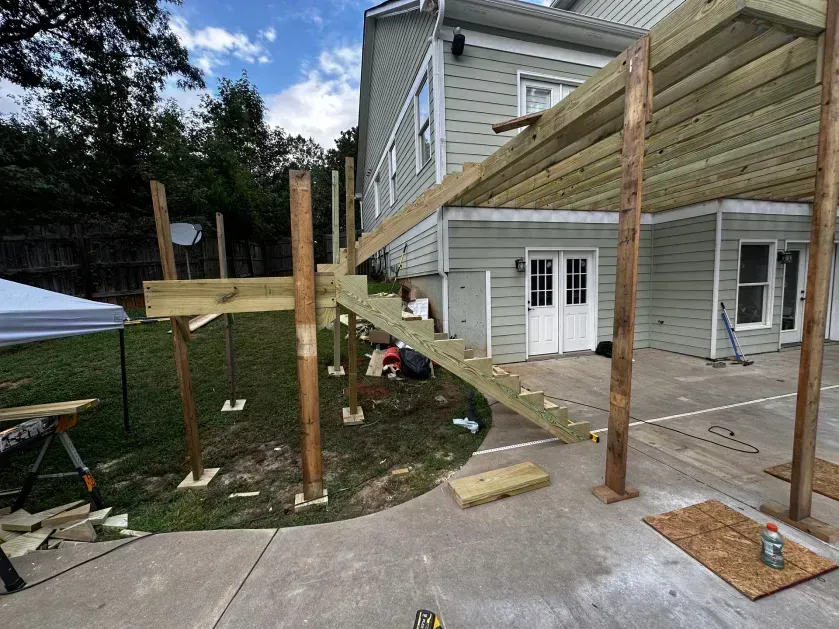 An outdoor view of a house under renovation with a wooden deck frame and stairs being constructed over a concrete patio.