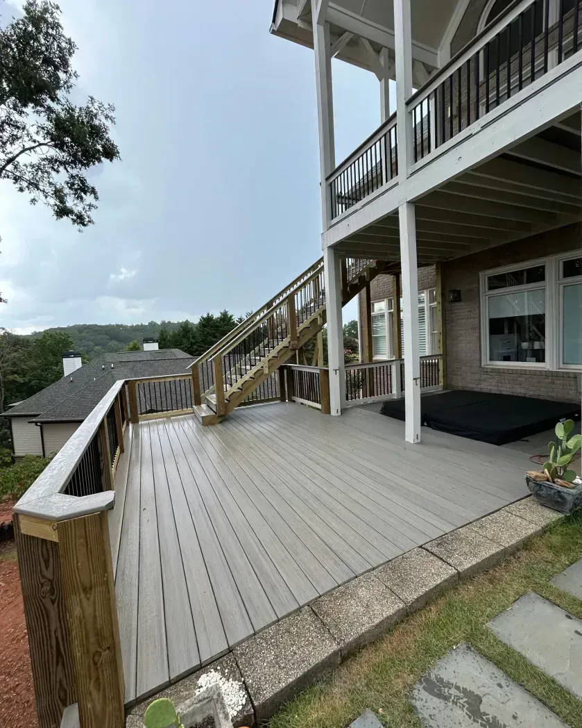 A two-story house exterior featuring a large grey composite deck, wooden stairs, and a black hot tub under the porch.