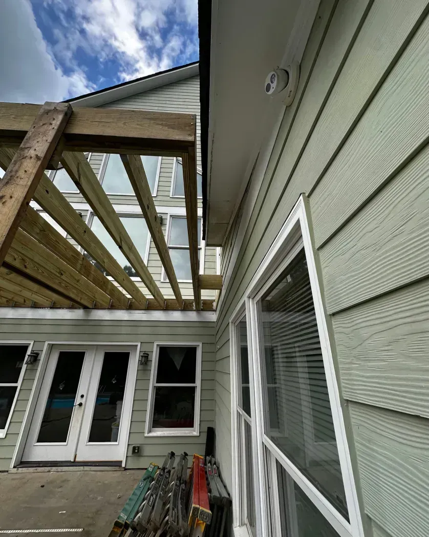 A low-angle view of a house exterior featuring light green siding, a white door, and an unfinished wooden pergola overhead.