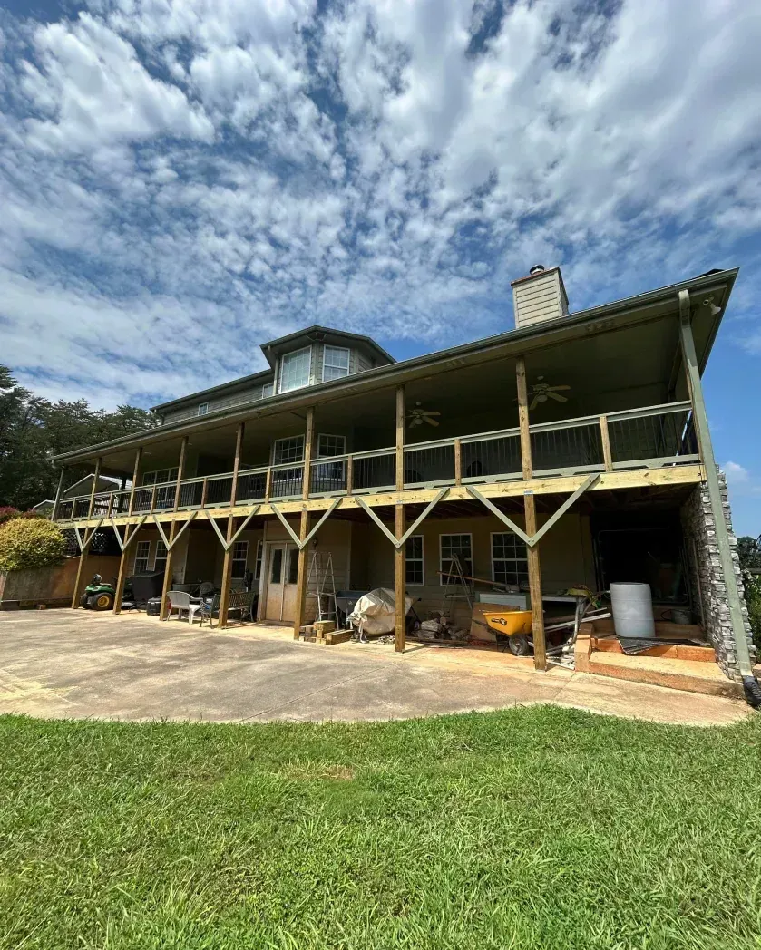 A two-story house with a large wooden wraparound deck, beige siding, a stone chimney, and a concrete patio area.