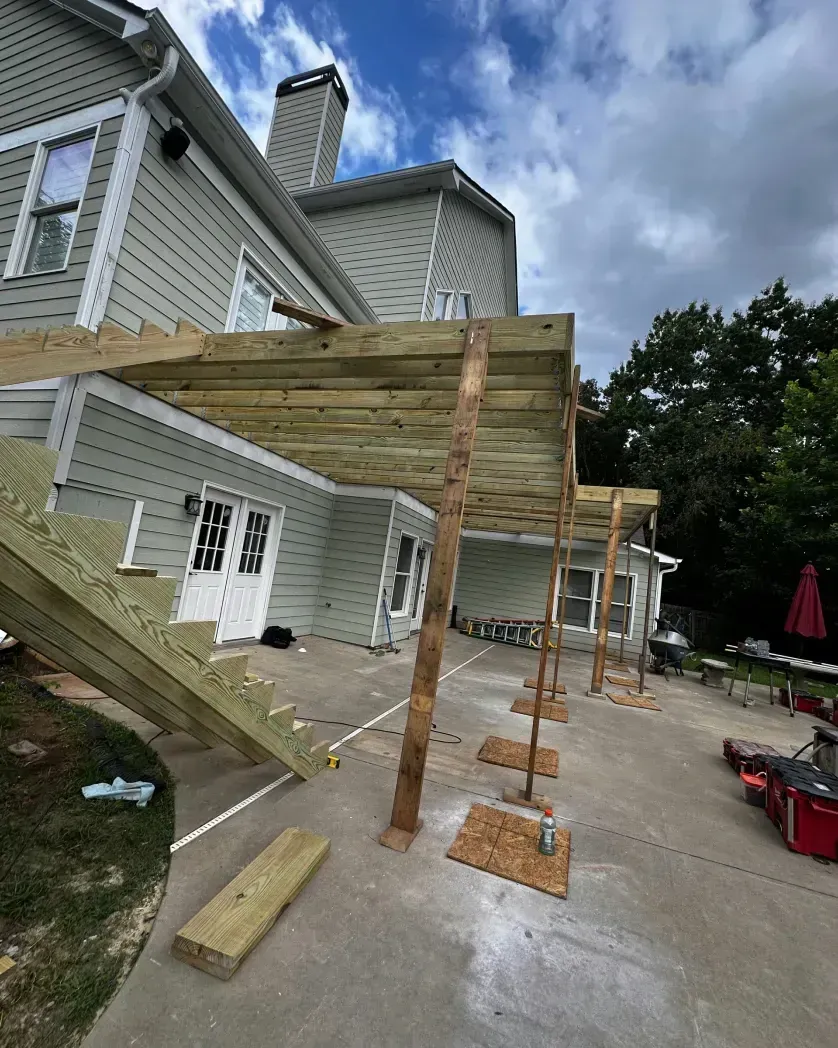 A low-angle view of a wooden deck frame under construction against the back of a house, supported by temporary poles.