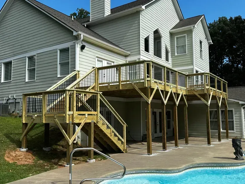 A large wooden deck with stairs attached to the back of a light gray two-story house overlooking a swimming pool.