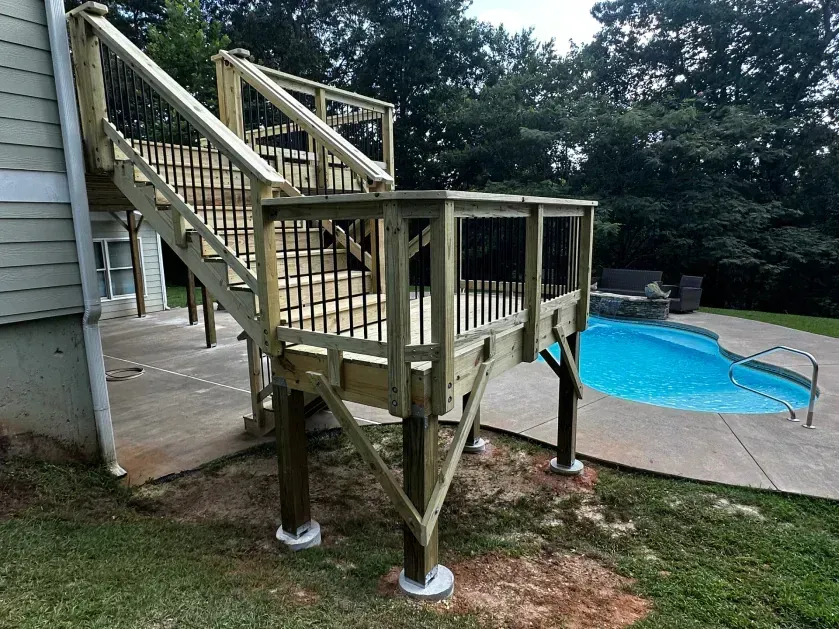 A wooden deck with stairs attached to a house, overlooking a swimming pool in a backyard setting.