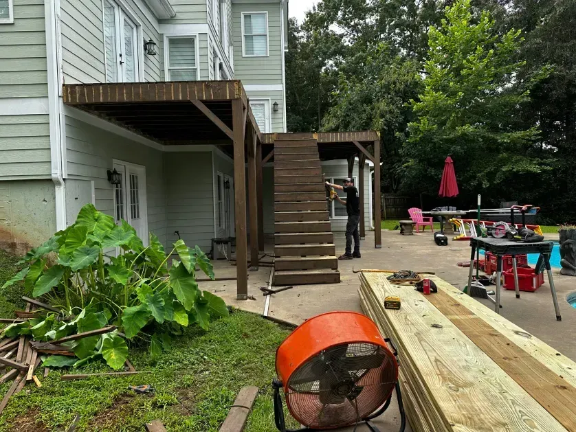 A person works on a wooden deck staircase under construction attached to a house next to a patio and swimming pool.