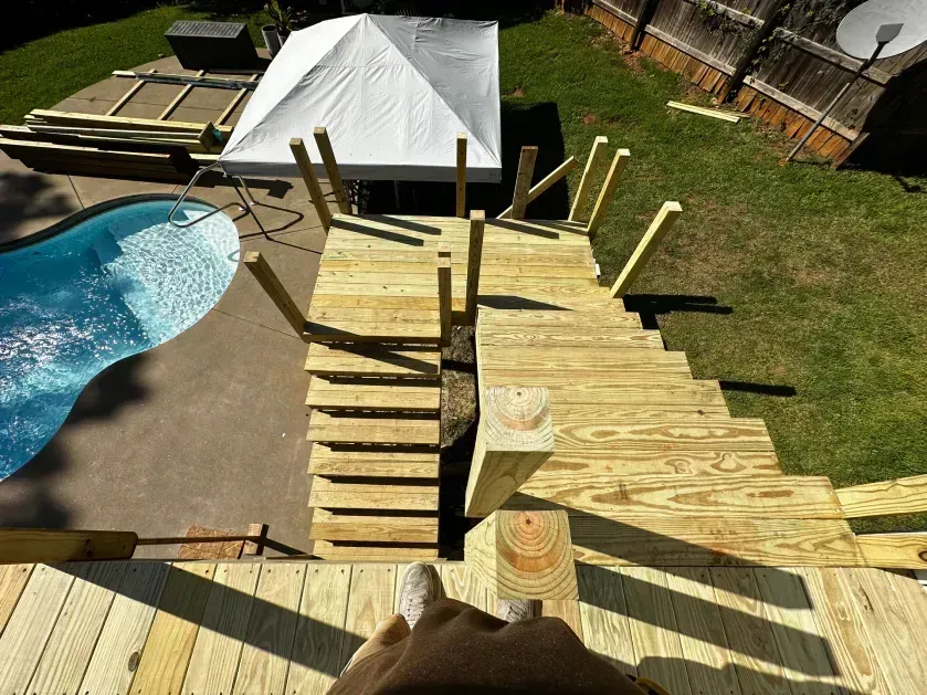 A first-person view looking down a newly built wooden deck staircase leading toward a swimming pool in a grassy backyard.