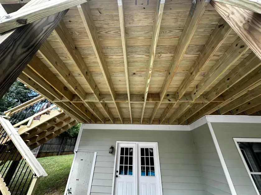 Underneath a wooden deck featuring exposed floor joists, with a staircase on the left and a double door on a house exterior.