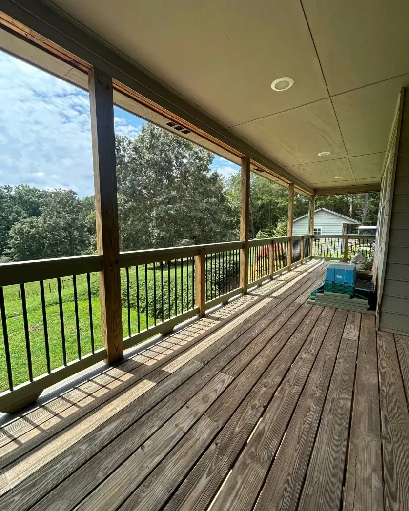 A covered outdoor wooden deck with black railings overlooking a grassy yard and trees under a blue sky.