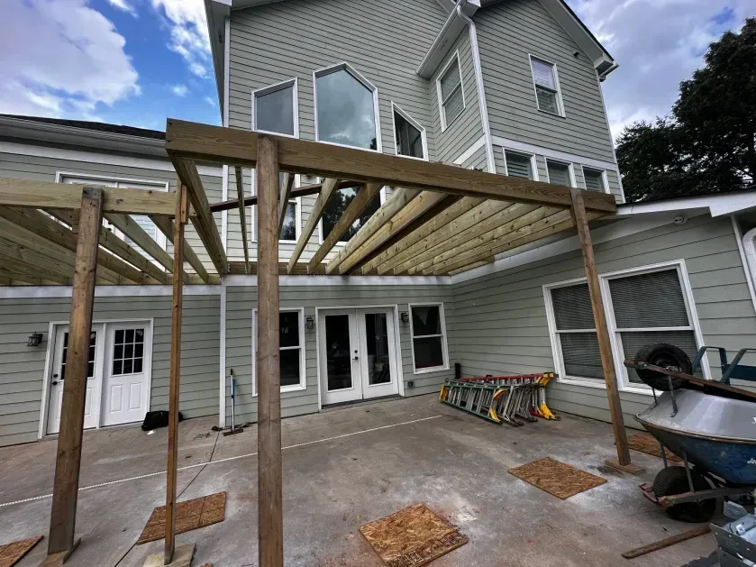 A wooden pergola frame under construction attached to the back of a light green house with a concrete patio.