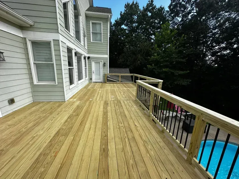 A newly built light-wood deck extends from a light-green house, overlooking a pool and trees.