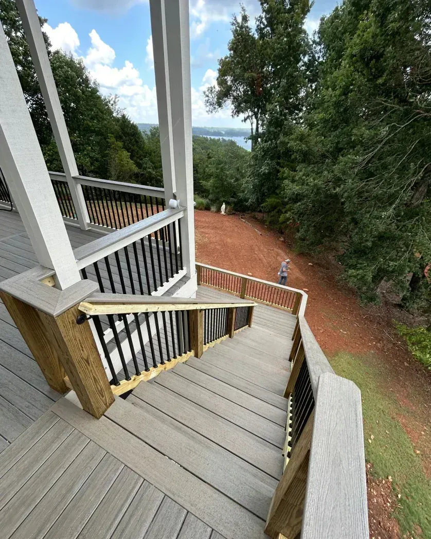 A wooden deck staircase leading down to a red-dirt yard and a distant lake, with a person standing on the slope below.