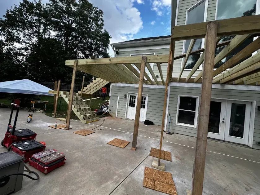 A wooden deck frame under construction extending from a house, with posts on a concrete patio and a staircase visible.