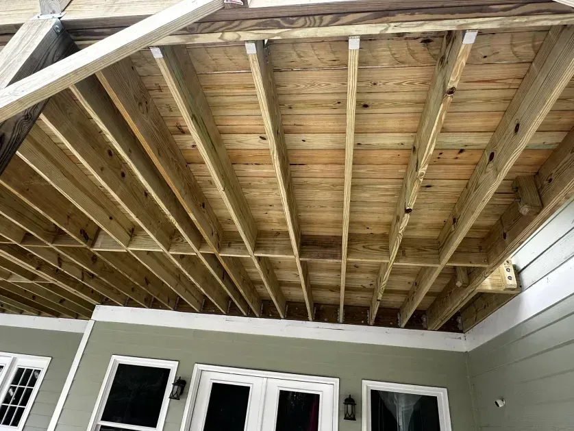 An upward view of a wooden deck frame with joists under construction, positioned above a house with green siding.