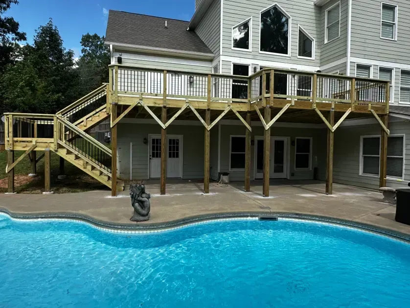 A large wooden deck with stairs overlooks a backyard swimming pool attached to a multi-story house.