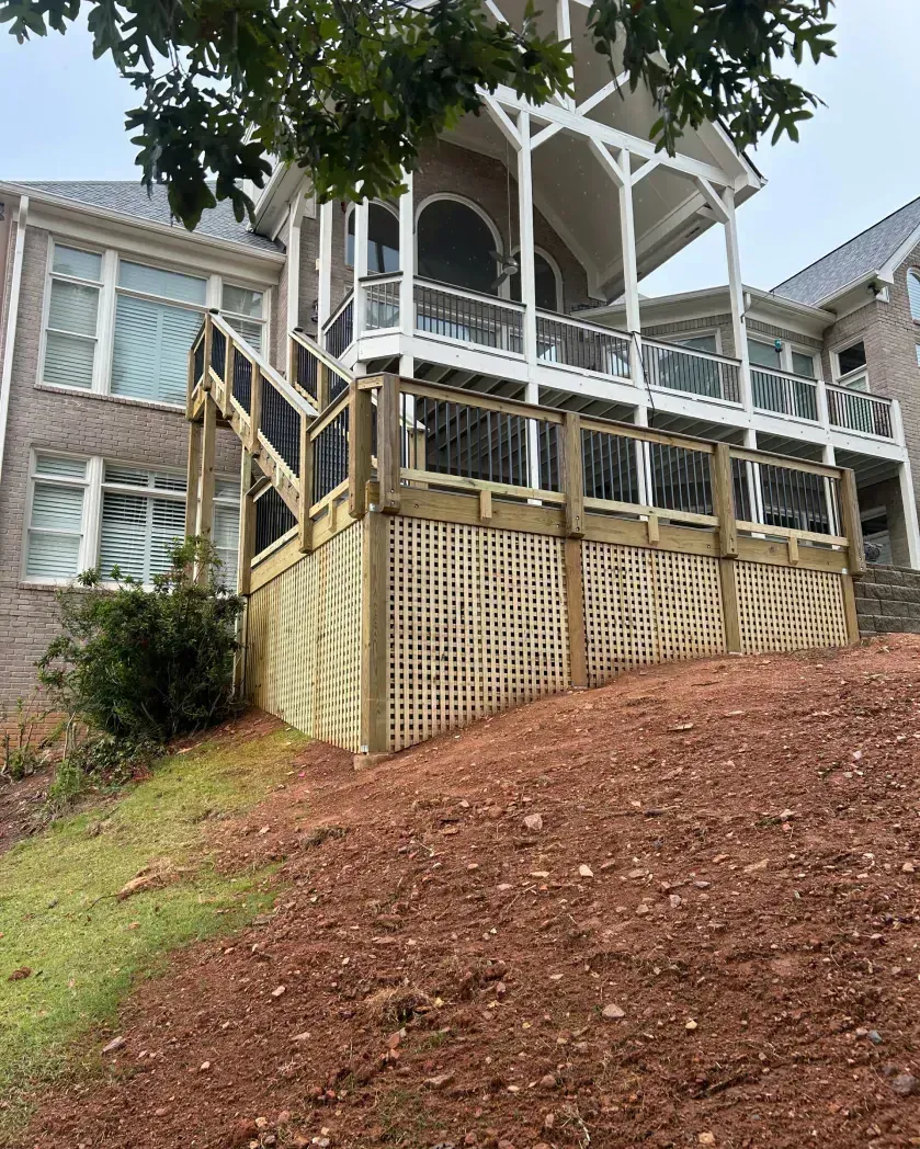 A tan brick house with a large wooden deck featuring white railings and lattice skirting on a sloped, mulch-covered yard.