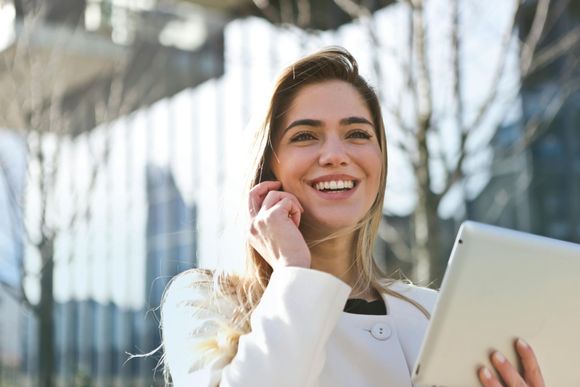 Woman in white jacket smiles while on the phone and holding a tablet outdoors in front of a building.