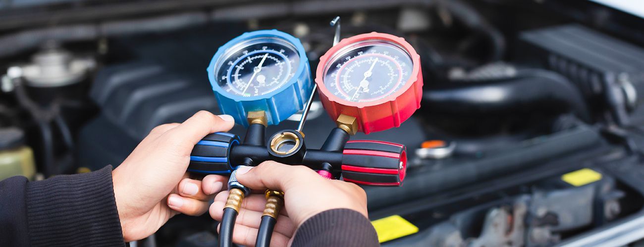 A pair of hands holds a manifold gauge set with blue and red dials over a car engine, used for testing AC systems.