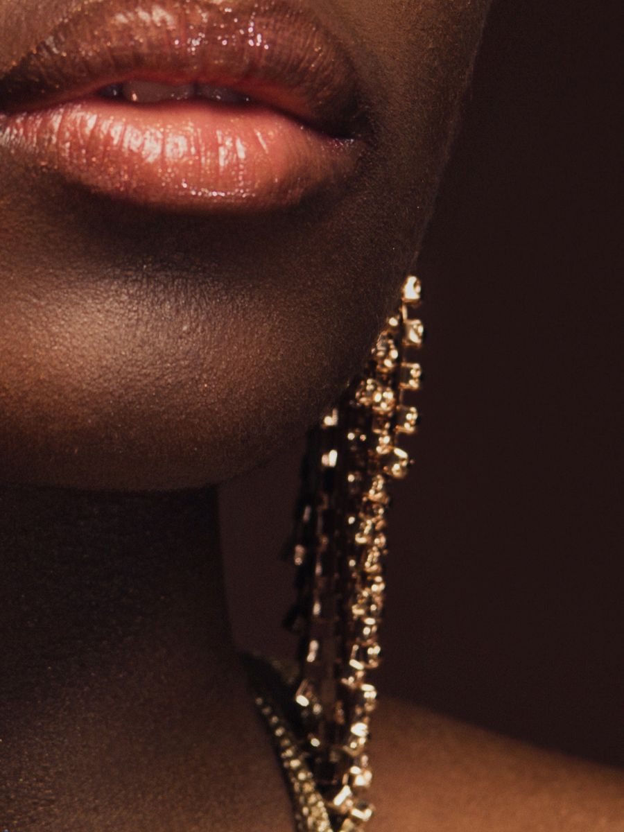 Close-up of person's lips and jaw; dark skin, glossy lip gloss, gold beaded earrings.
