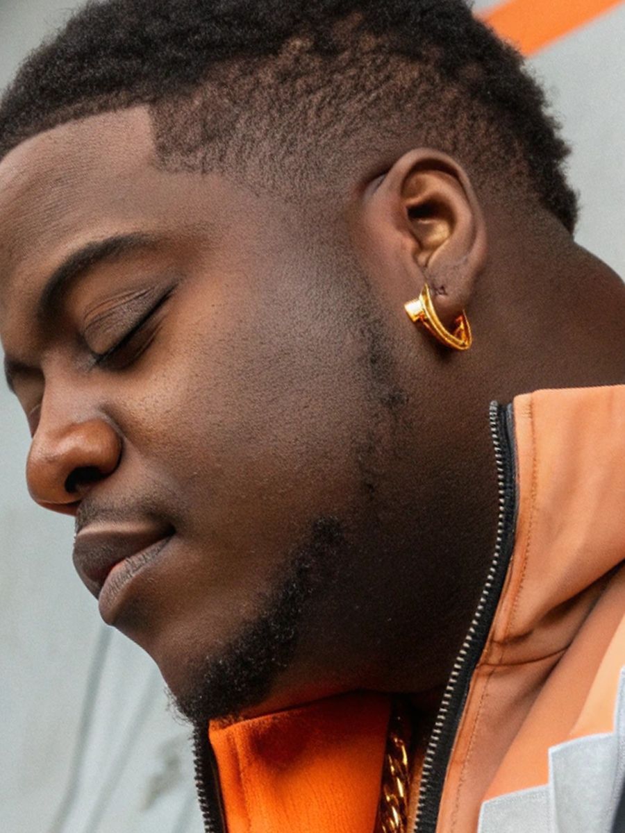 Man with closed eyes wearing gold earrings, gold chain, and orange jacket.