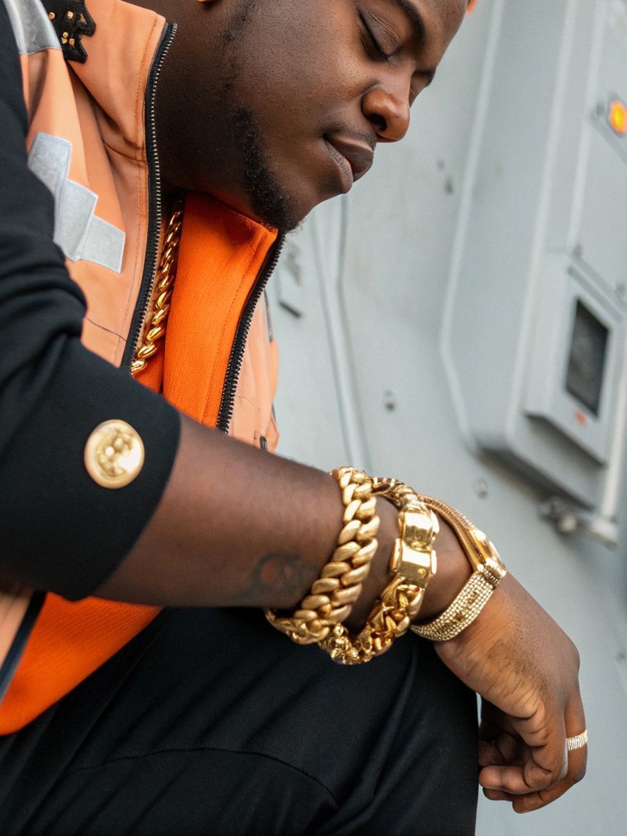 Man wearing multiple gold chains and bracelets, in an orange vest and a military setting.