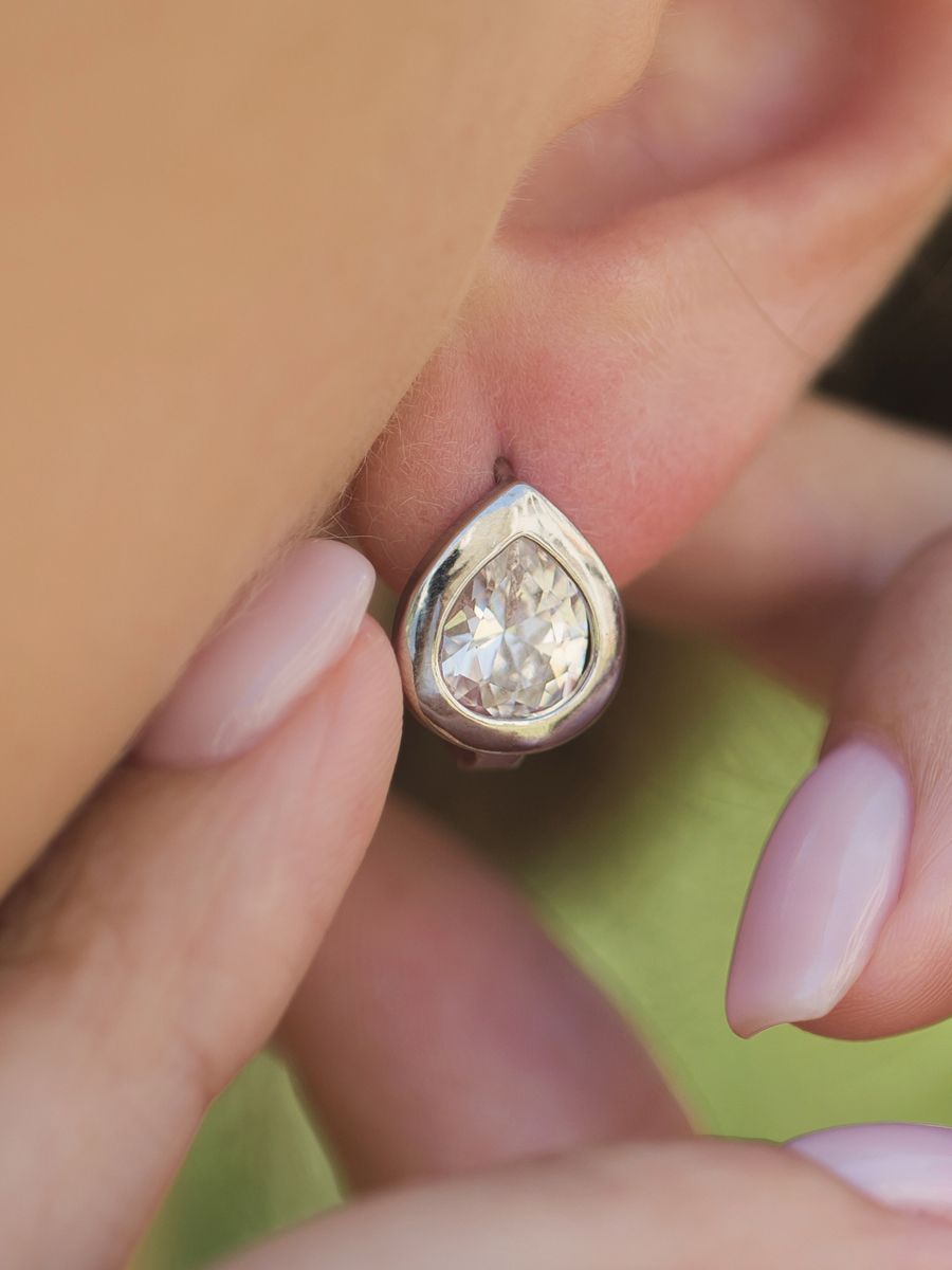 Close-up of a person's ear with a teardrop-shaped diamond earring.  A hand gently touches the earring.