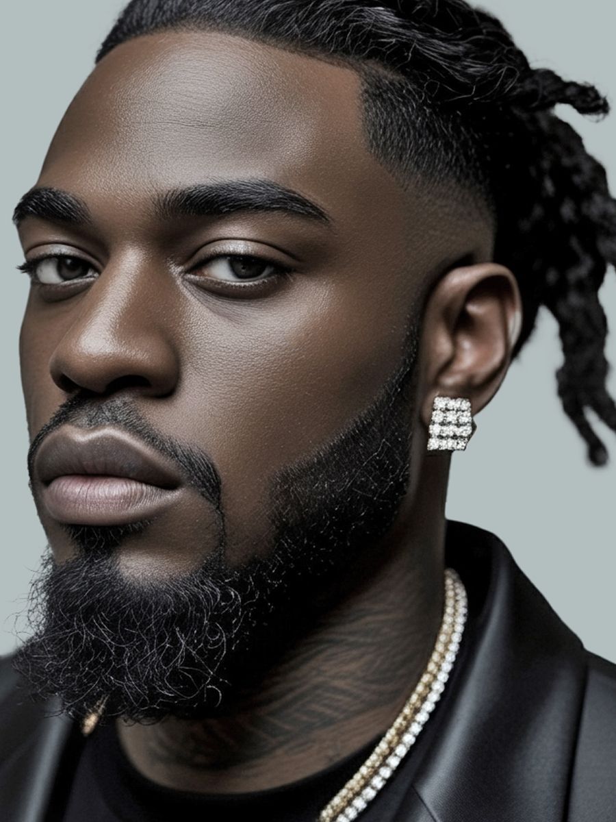 Man with dark skin, wearing a black jacket, beard, and gold jewelry. He gazes at the viewer.