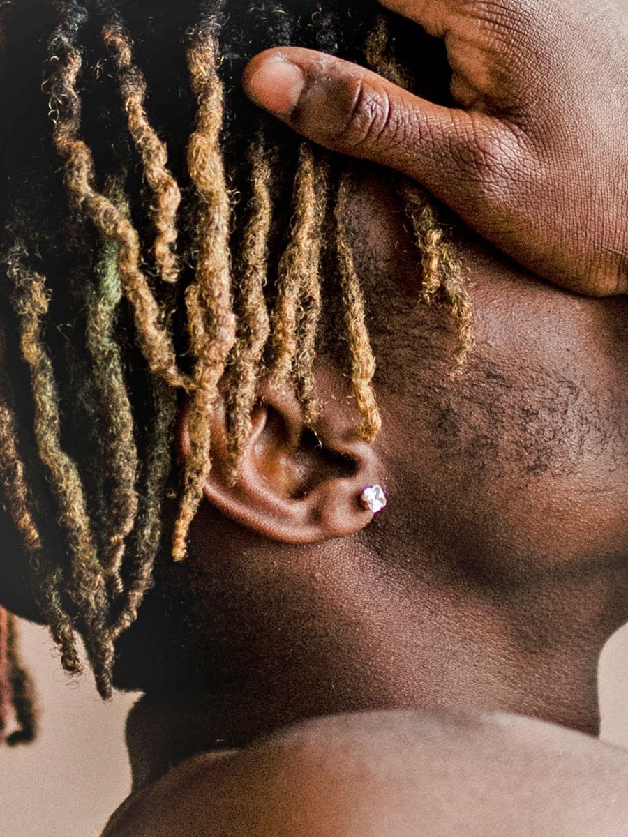 Person with blonde and black dreadlocks, hand on head, and earring.