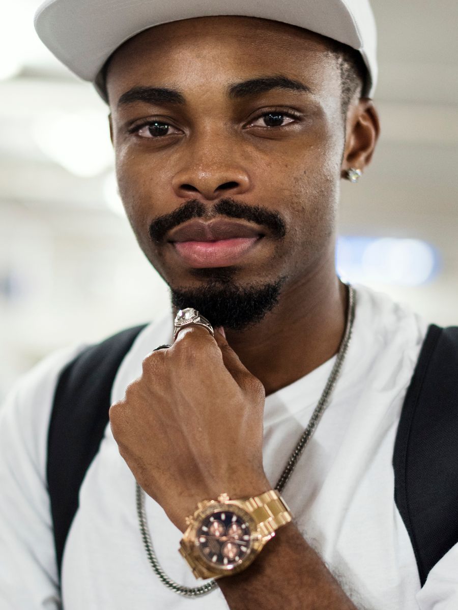 Man with a goatee, wearing a white cap, watch, and silver chain, resting his chin on his hand.