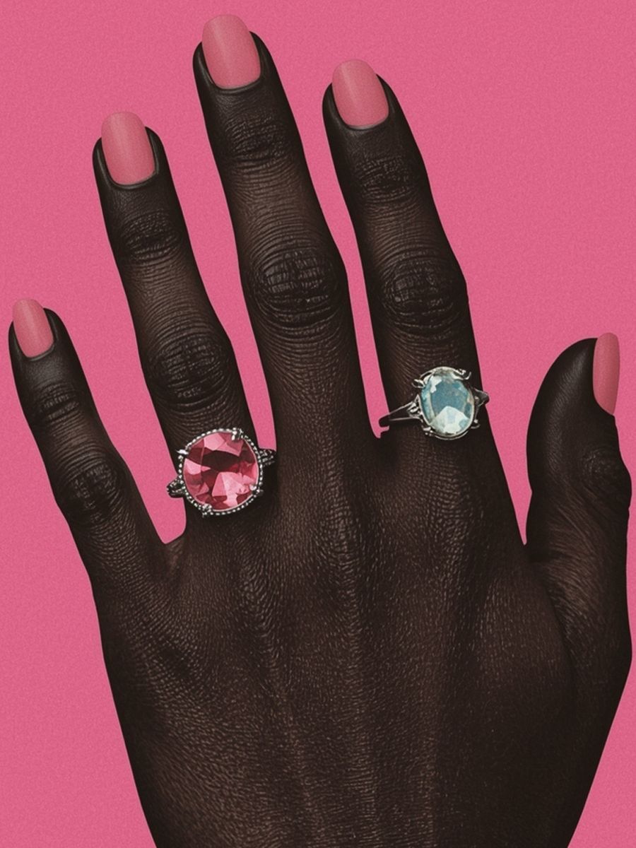 Dark-skinned hand with pink nails wearing two jewel-toned rings against a pink backdrop.