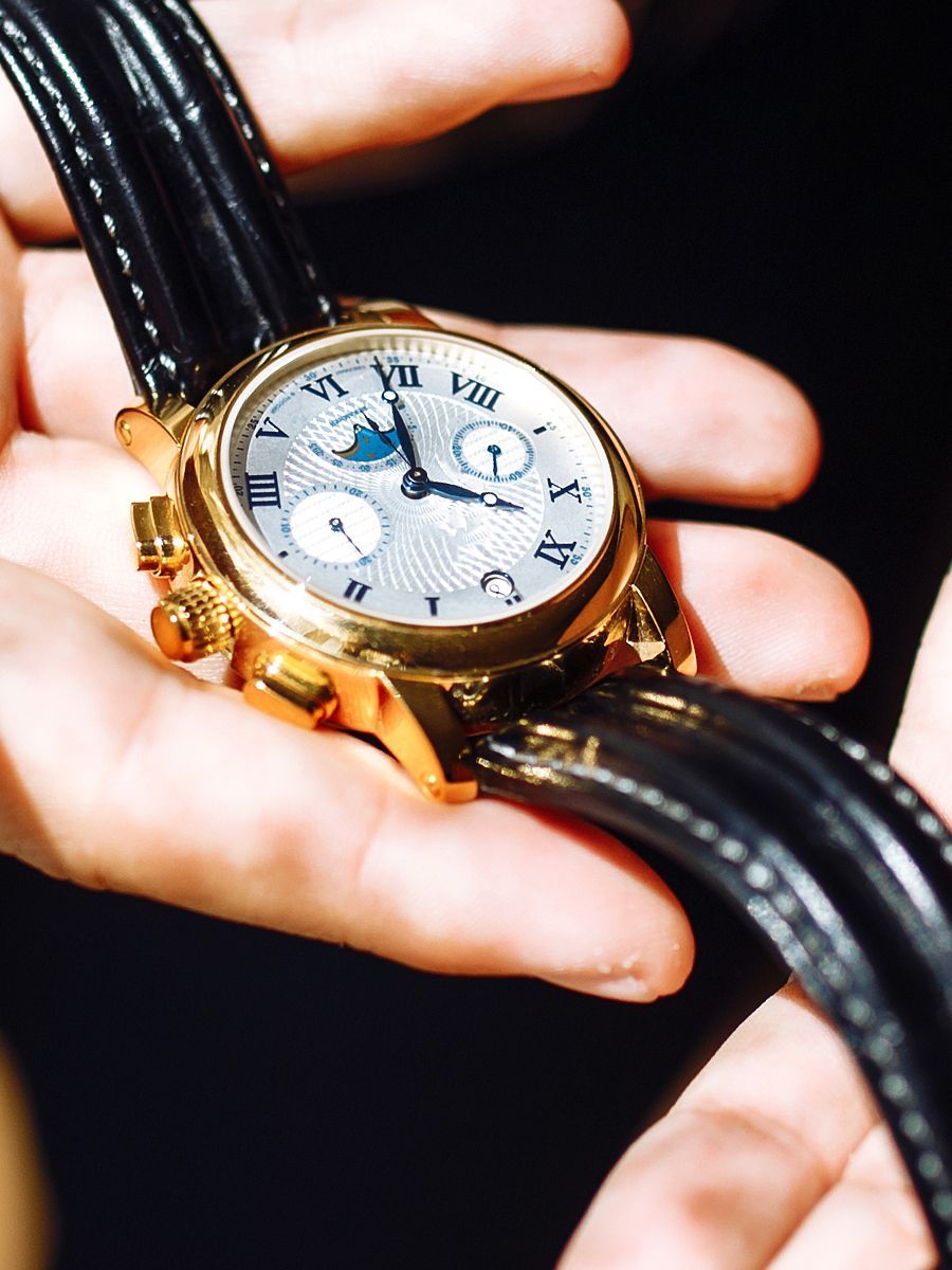 Gold wristwatch held in hands, featuring a light-colored face with subdials and a black leather strap.