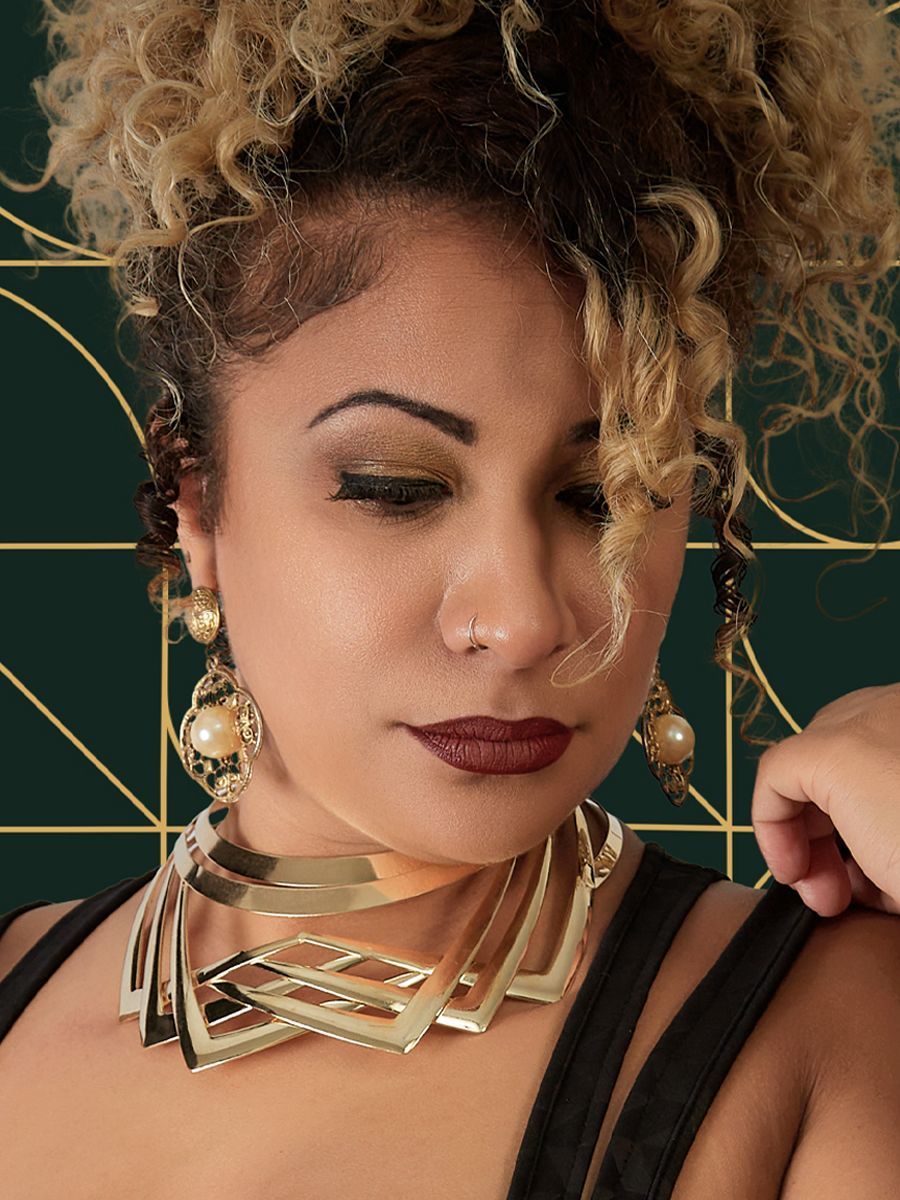 Woman with curly blonde hair, gold jewelry, and dark makeup, looking down.