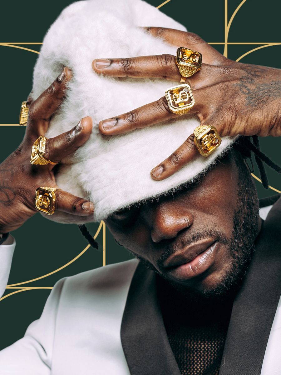 Man wearing a white fuzzy hat, many gold rings, white suit jacket, dark skin.