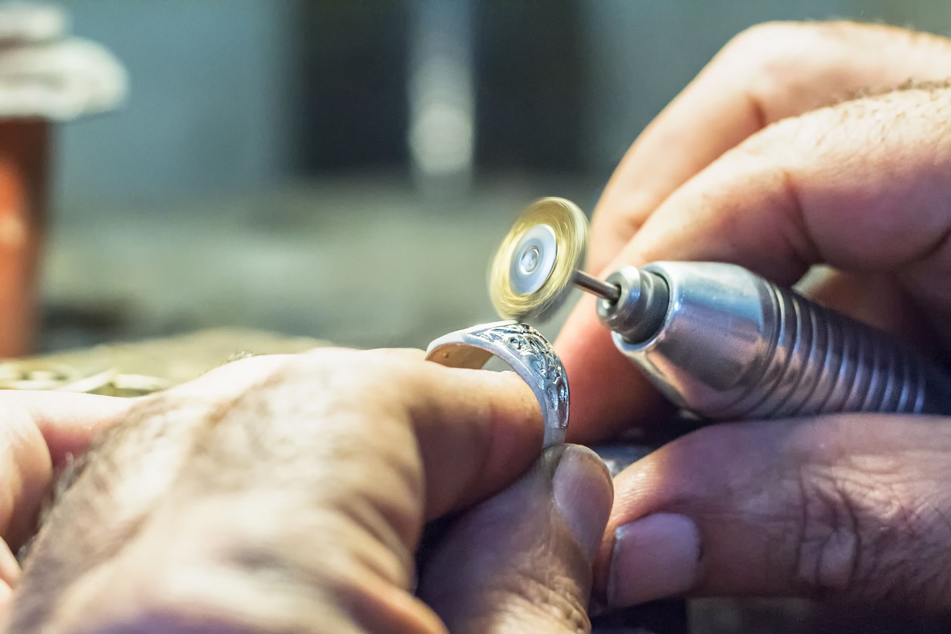 Jeweler uses a rotary tool to polish a silver ring with inlaid gemstones. Close-up of hands at work.