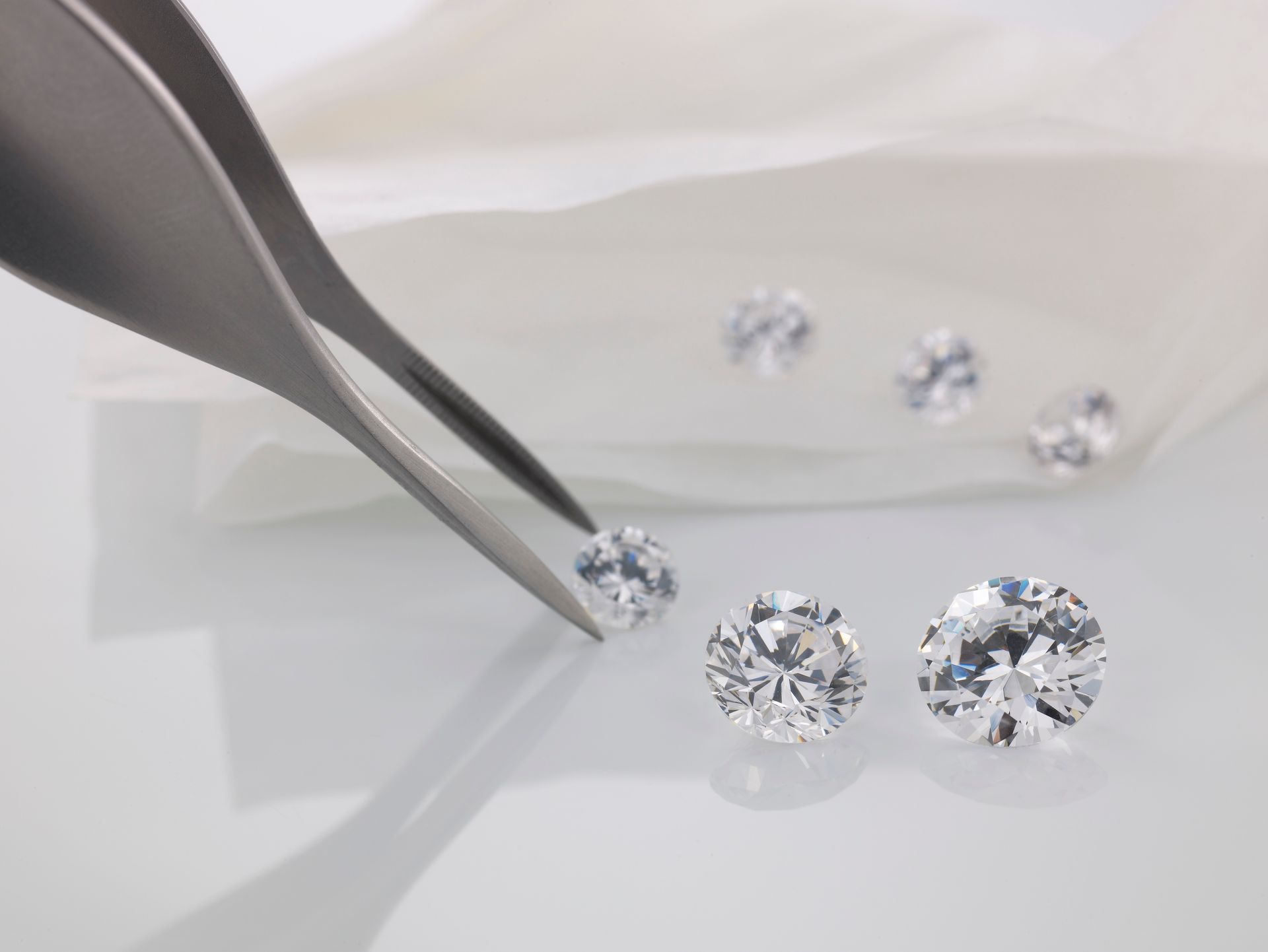 Tweezers holding a diamond, several loose diamonds on white surface.
