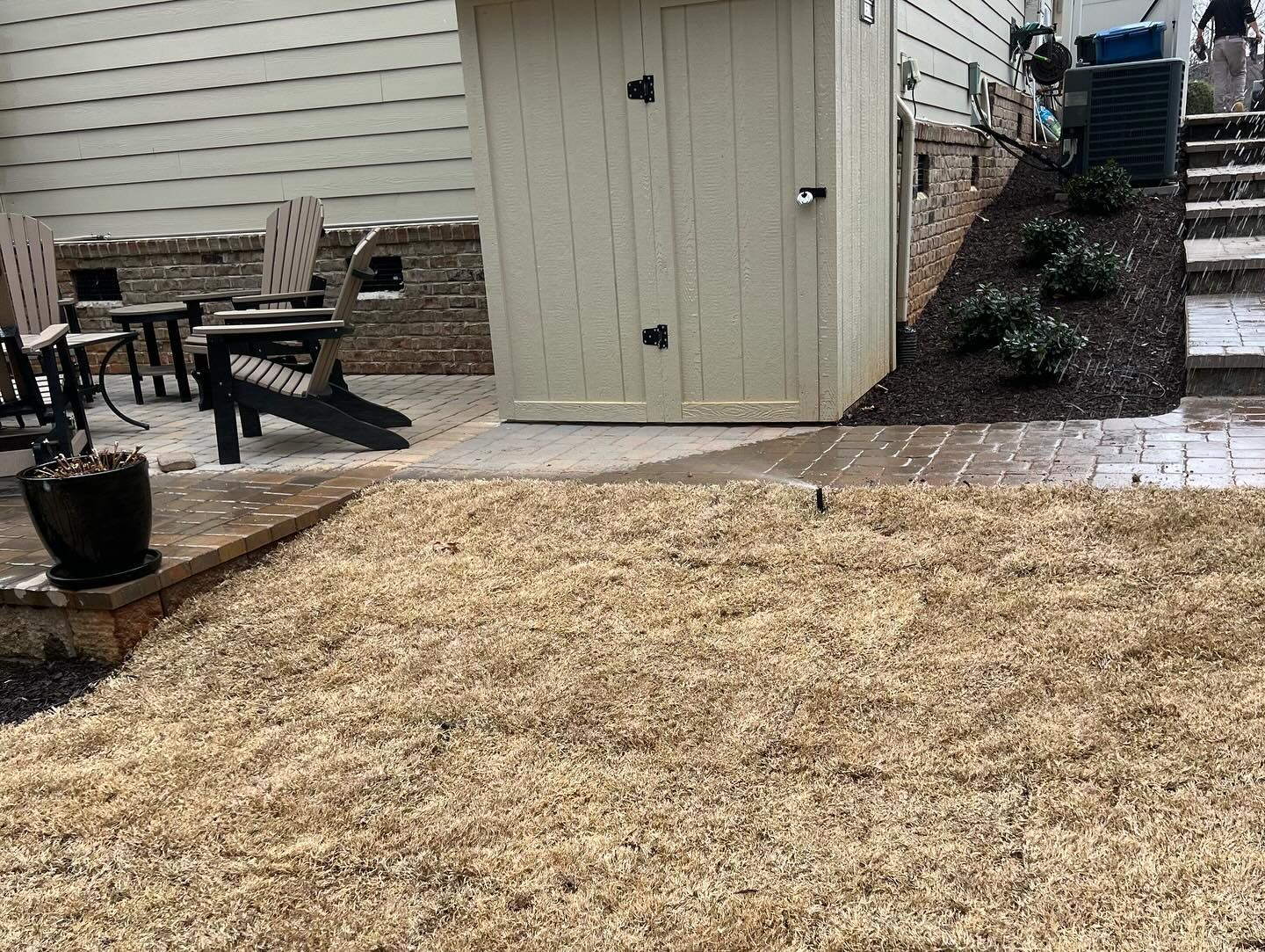 Backyard with beige shed, patio furniture, dry grass, and brick walkway.