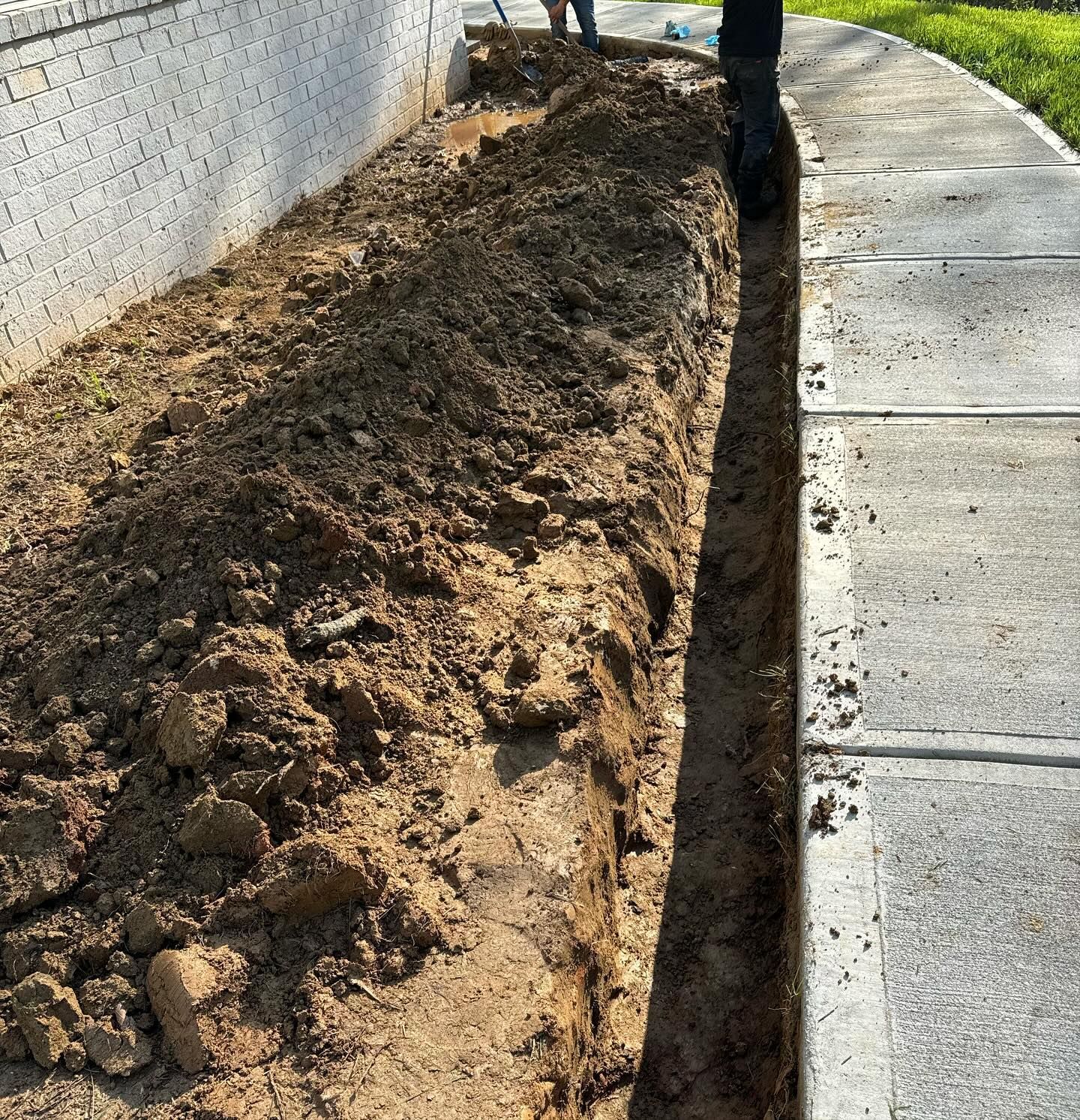A trench dug in brown earth next to a sidewalk and a white brick wall. Someone is standing over the trench.