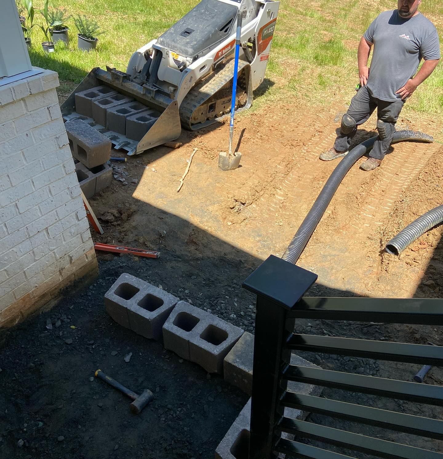 Construction site: Man watches a small excavator laying cinder blocks near a building; a hammer is on the ground.