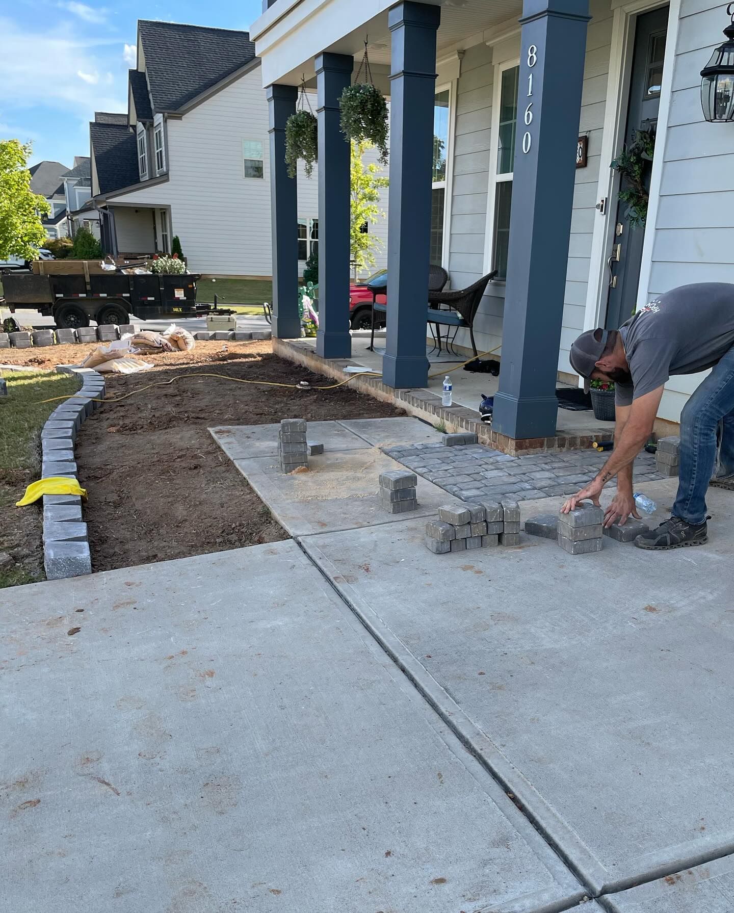Man laying paving stones on a porch. Grey blocks, blue columns, tan soil.