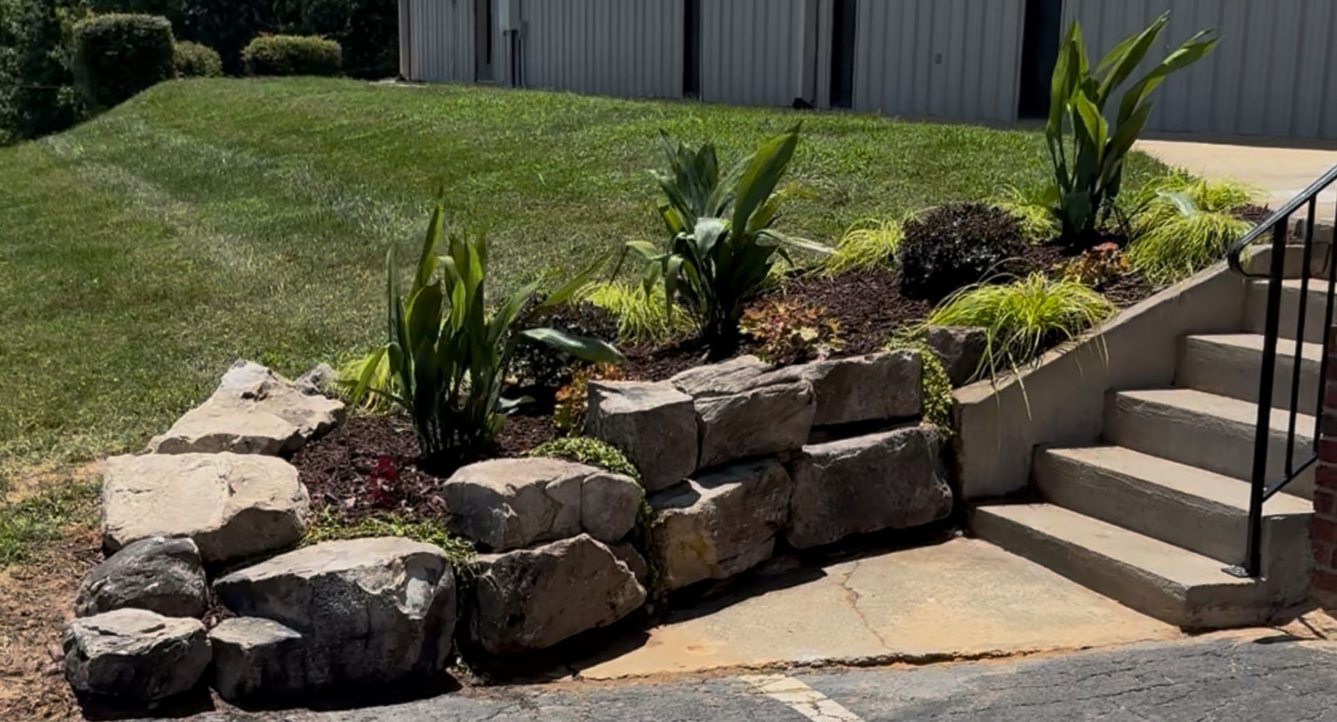 Stone retaining wall with planted flowers and shrubs next to concrete steps leading up to a building.