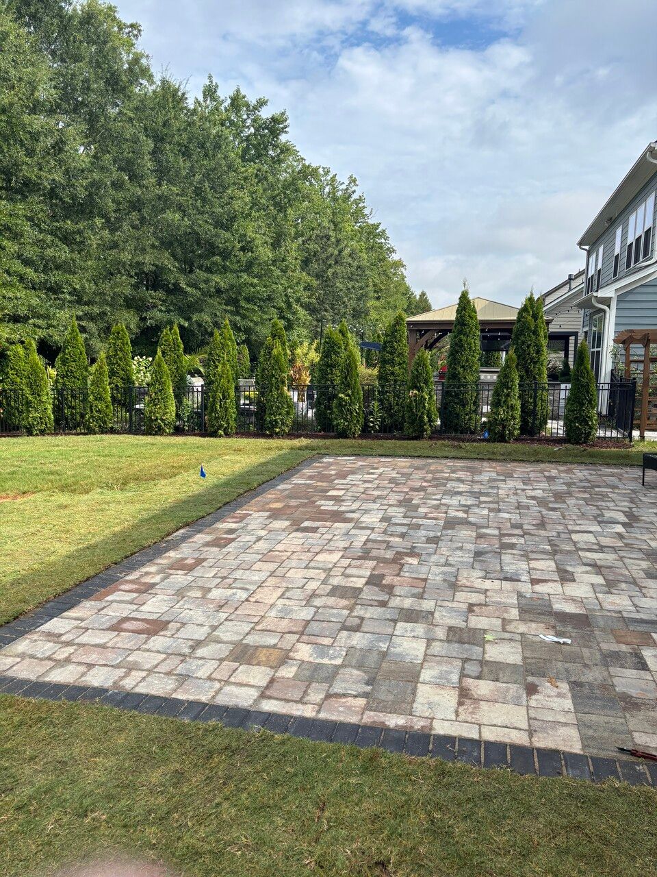 Brick patio with bordering green grass, lined by evergreen trees and a house under a cloudy sky.