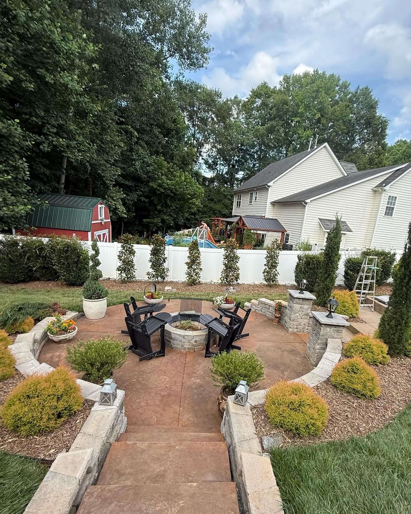 Patio with fire pit and Adirondack chairs, surrounded by landscaping and a white fence.