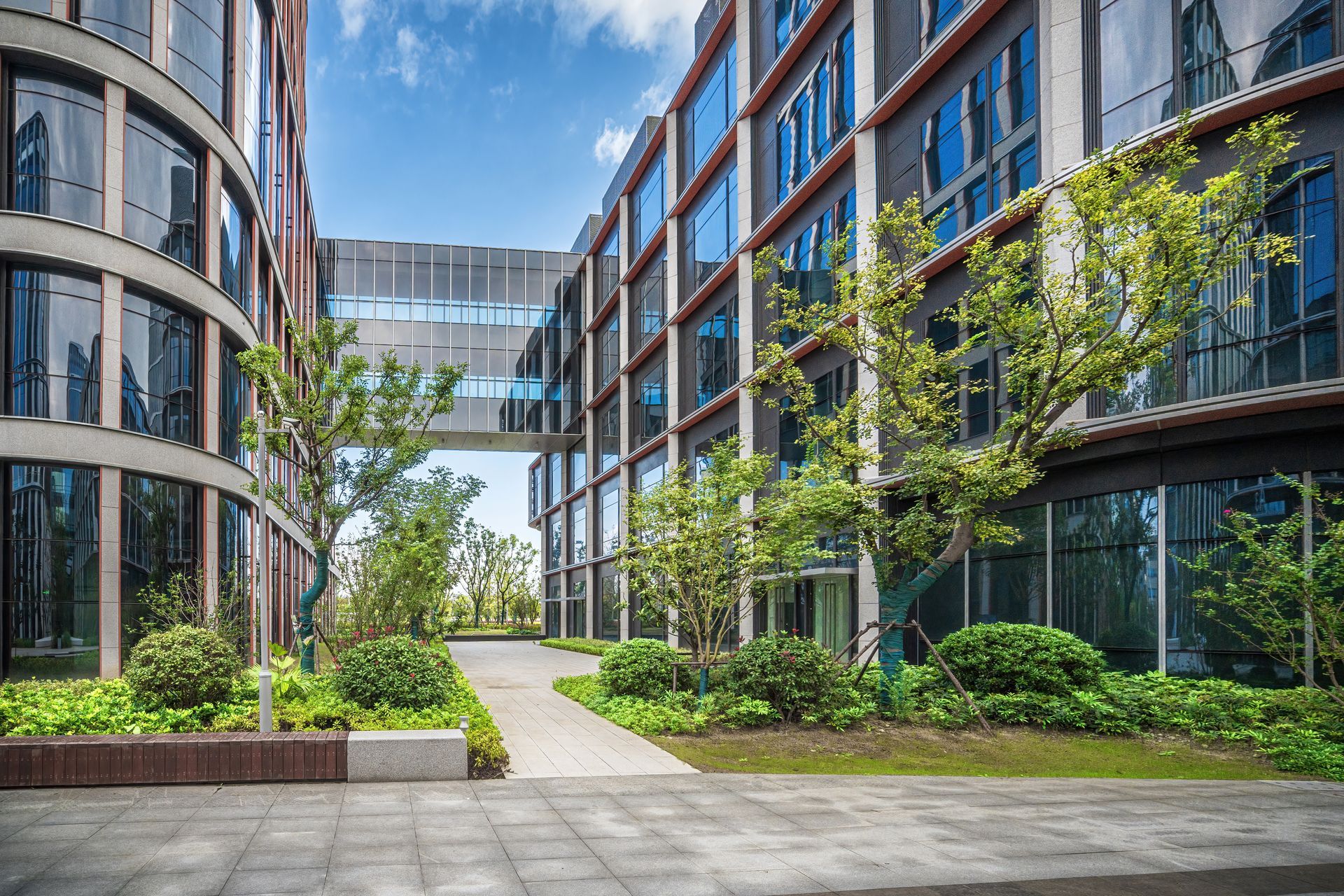 Modern office buildings with a walkway between them, landscaped courtyard, sunny day.