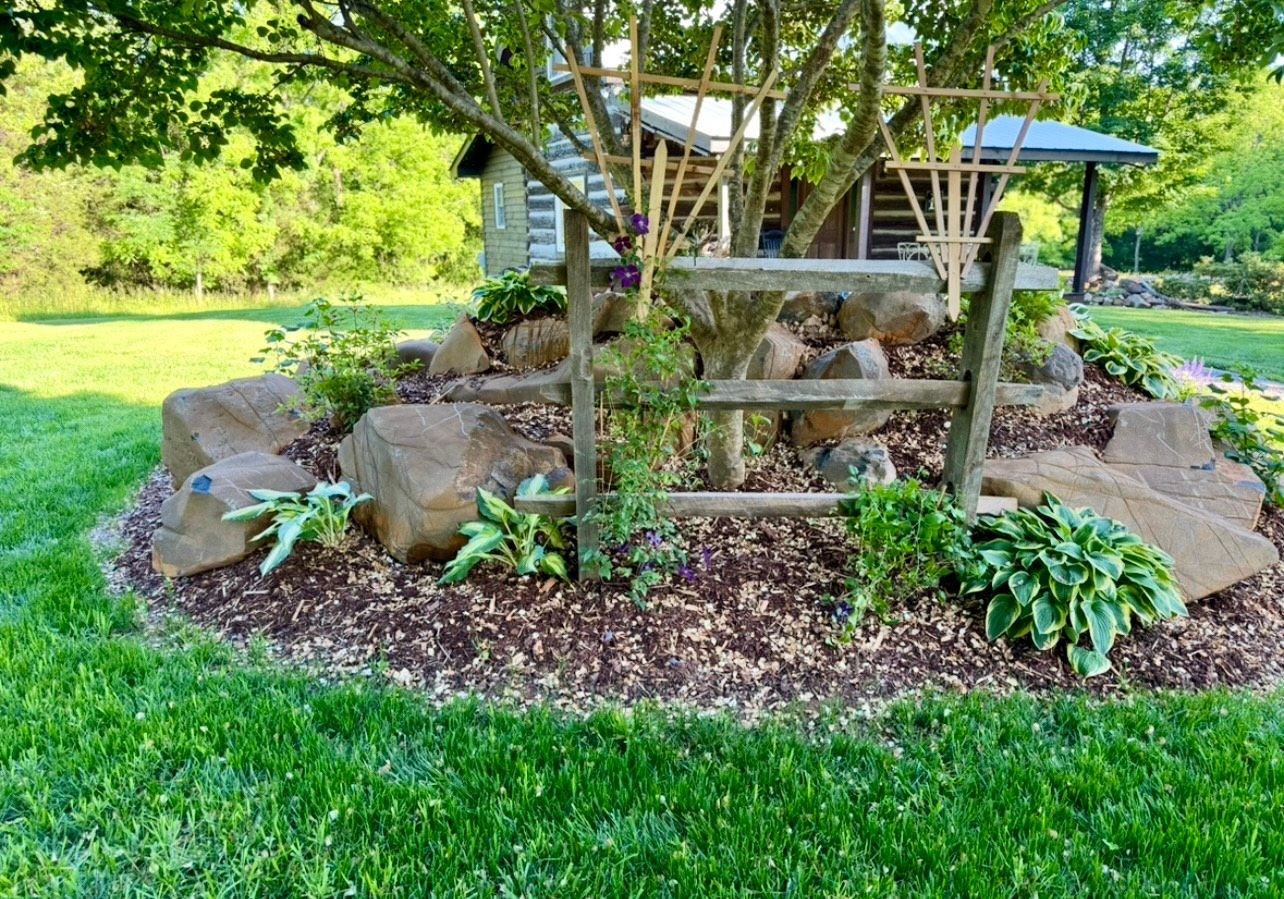 Brick path winds through a landscaped garden with a bench and small blue building in the background, surrounded by trees.