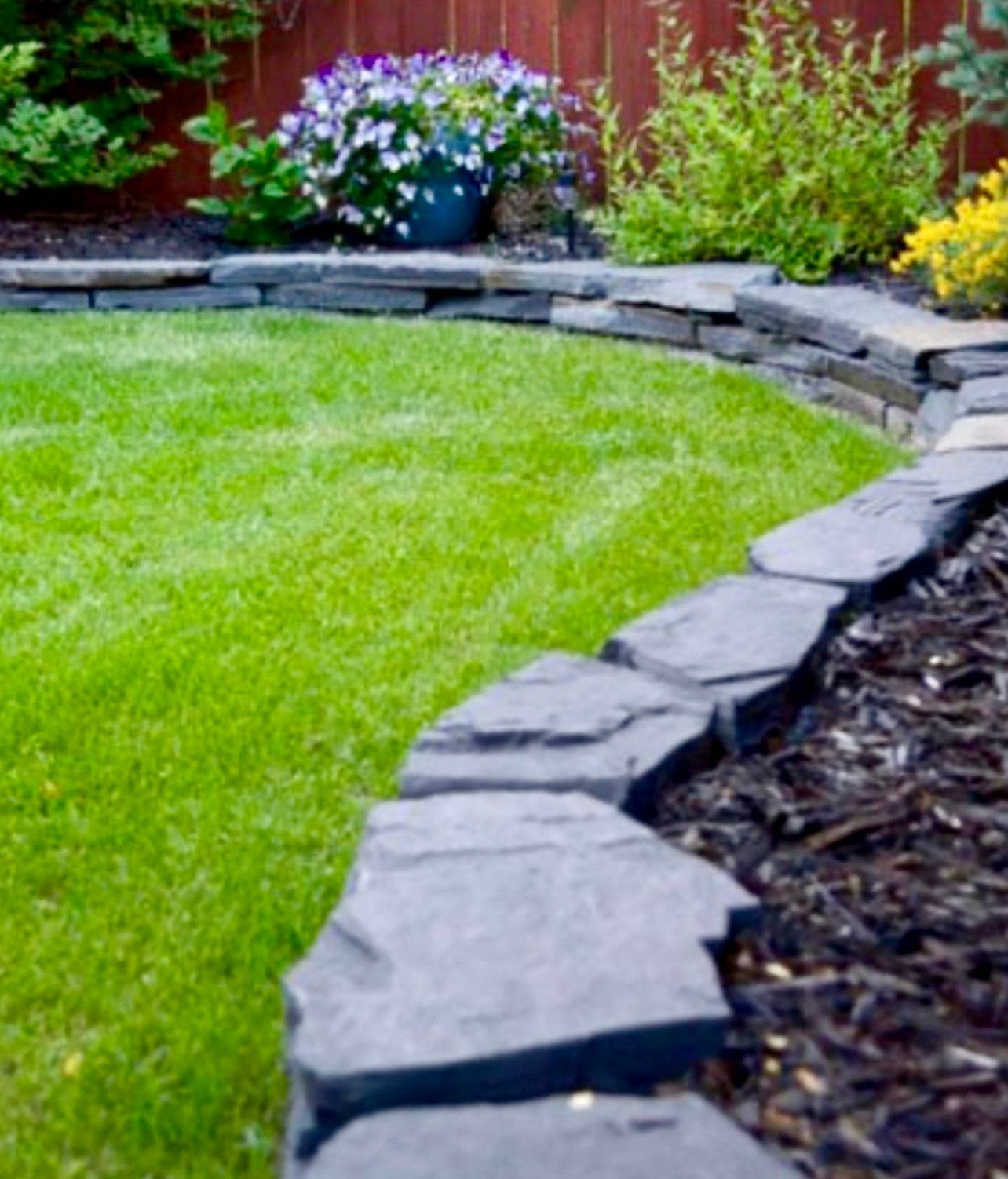 Lush green lawn bordered by dark stone, with flowers and mulch in the background.