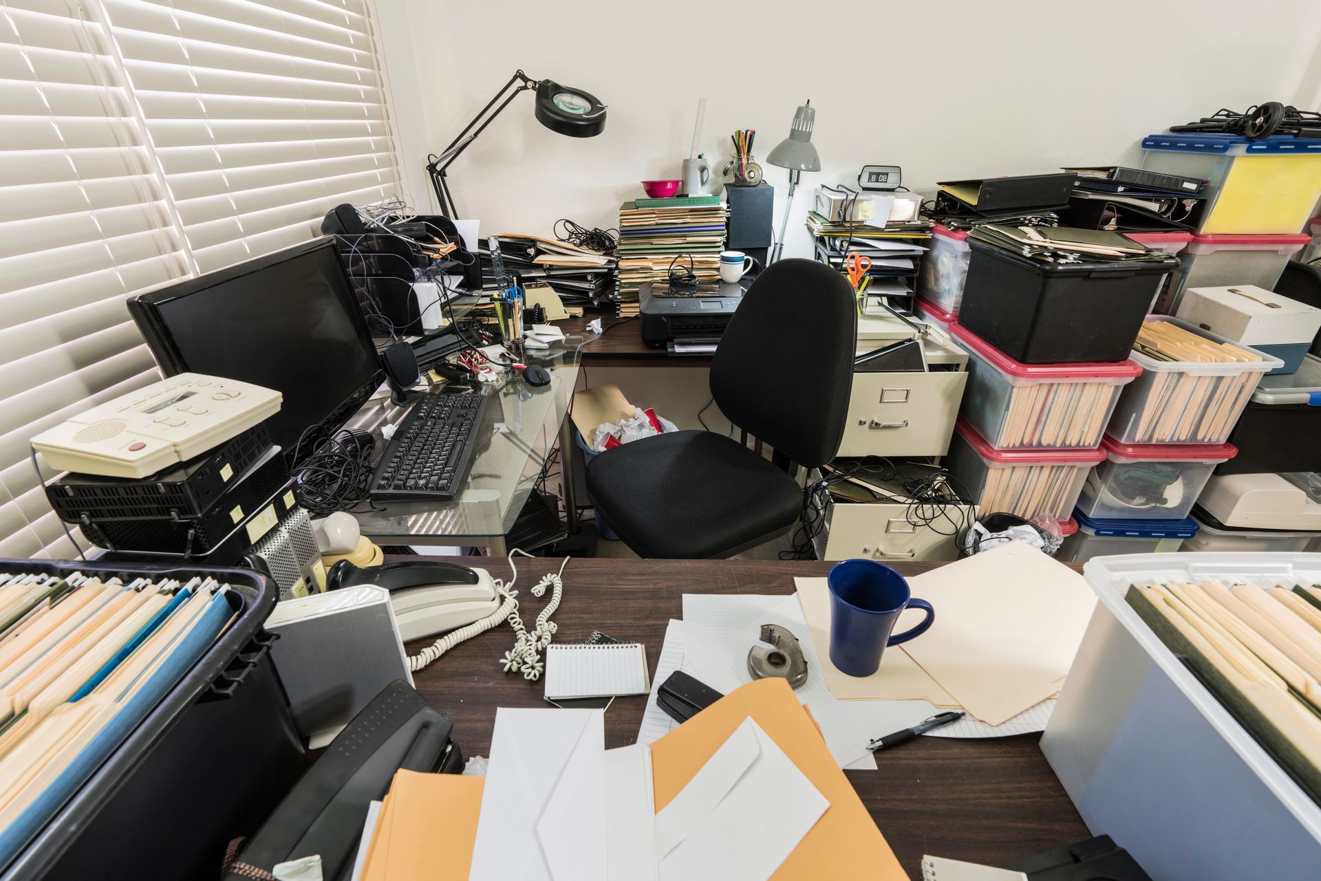 Cluttered desk with computer, papers, and office supplies; a person is seated in a chair.