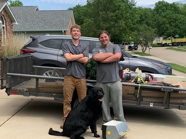 Two men and a dog are standing in front of a trailer.