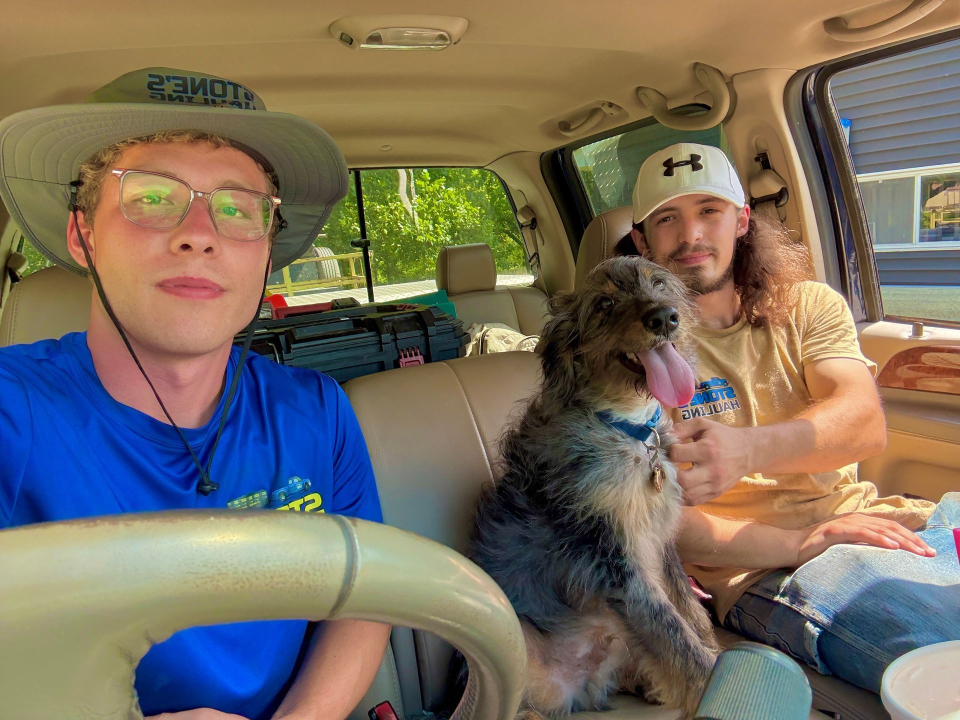 Two men and a dog in a car, smiling at camera. One man wears a hat and glasses, the other a cap. Sunny outdoor setting.